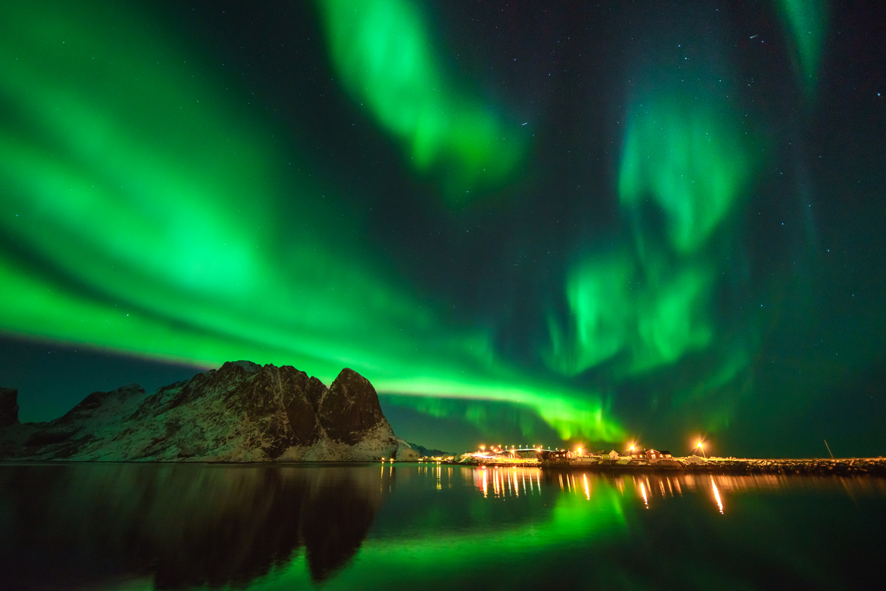 Aurora boreală verde strălucitoare luminând cerul nopții deasupra unui munte înzăpezit și reflectându-se în apele calme, in luminile unei mici așezări rurale în fundal.