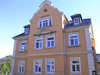 Hell gelbes, historisches Wohngebäude mit vielen Fenstern und dekorativem Giebel vor blauem Himmel.