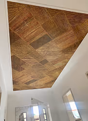 Decke mit einem dekorativen Holzpaneel in verschiedenen Mustern in einem hellen Eingangsbereich mit Fenstern und weiß gestrichenen Wänden.