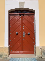Rote doppelte Holztür mit silbernem Türgriff und dekorativen Nieten in einem gelben Gebäude.