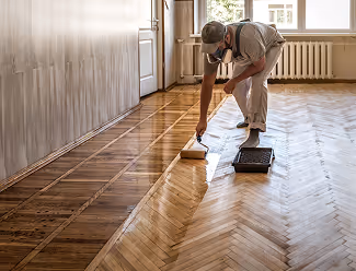 Mann in Arbeitskleidung trägt Parkettboden mit Lackrolle in einem hellen Raum auf.