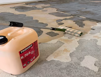 Kanister mit Aufkleber 'Thomsit R 766' neben einer gerollten Rolle auf einem Boden mit Flecken und Ausbesserungen.
