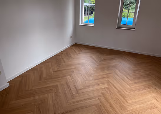 Leerer Raum mit weißen Wänden und zwei Fenstern, Sicht auf einen hellbraunen Fischgrätenparkettboden.