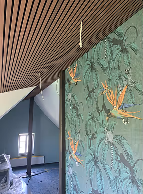 Raum mit schrägem Holzbalken an der Decke, grünem Tapeten mit tropischem Vogel- und Pflanzenmuster und offenem Fenster.