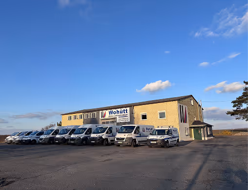 Gelbes Bürogebäude mit dem Schild 'Wohütt' und einer Reihe weißer Firmenfahrzeuge auf einem Parkplatz unter blauem Himmel.
