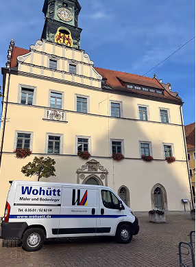 Weißer Kleintransporter mit Werbung für Maler und Bodenleger vor einem historischen Gebäude mit Turmuhr und blauer Himmel.