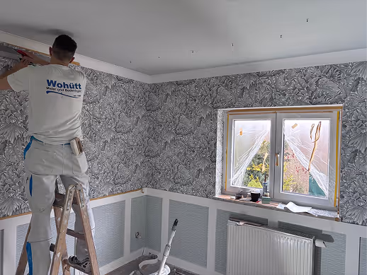 Ein Mann auf einer Leiter bringt Tapetenoberkante in einem Zimmer mit gemusterten Wänden an.