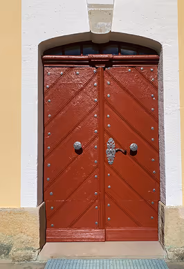 Rotes hölzernes Doppeltür mit Metallbeschlägen und weißem Steinrahmen vor gelber Wand.