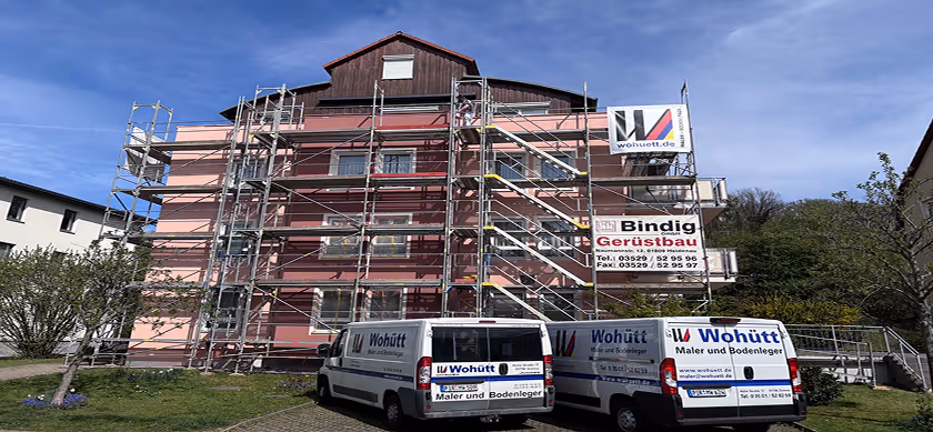 Mehrstöckiges Wohnhaus mit rosa Fassade, umgeben von einem Gerüst, vor dem zwei weiße Firmenfahrzeuge von Wohütt Maler und Bodenleger parken.
