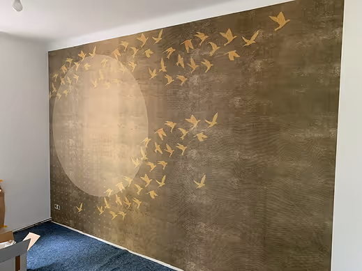 Wand mit dunklem Muster und goldenem Design von Vögeln, die um einen großen, hellen Kreis fliegen.