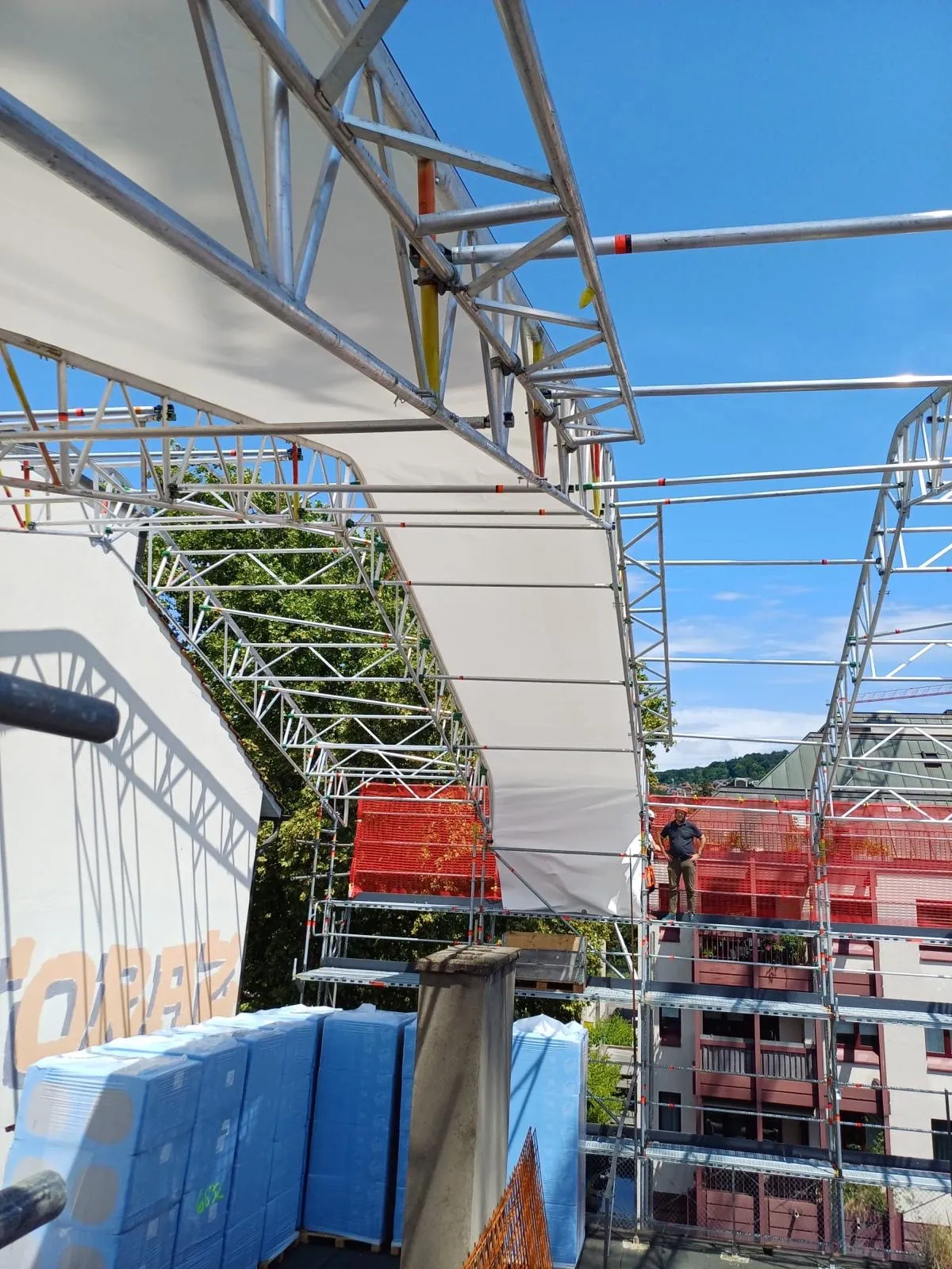 Stillasbygning med hvit duk spent over metallrammer på en byggeplass, en mann står på stillaset ved siden av en bygning med røde balkonger under en klar blå himmel.