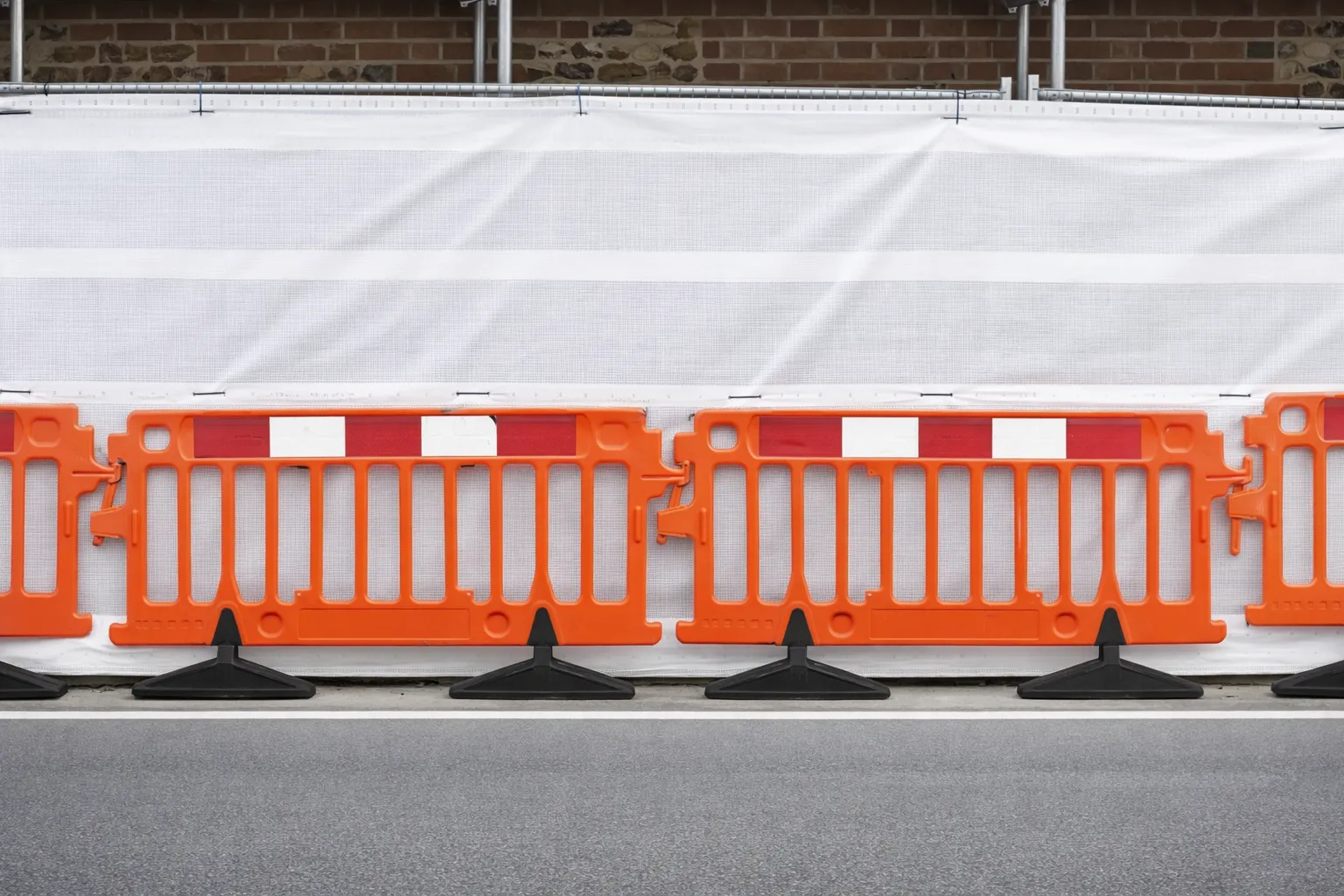 Orange plastic safety barriers linked together on a street in front of a white mesh construction fence with a brick wall behind it.