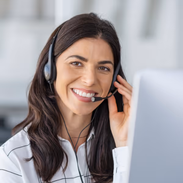 Lächelnde Kundendienstmitarbeiterin mit langem dunklem Haar und Headset vor einem Computerbildschirm.