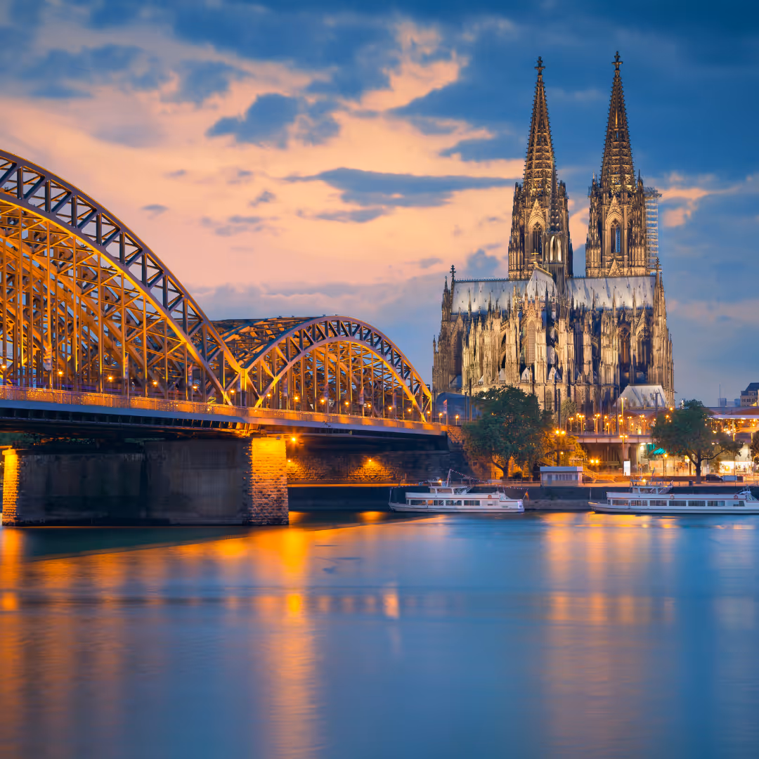 Die beleuchtete Hohenzollernbrücke über den Rhein mit dem Kölner Dom im Hintergrund bei Sonnenuntergang.
