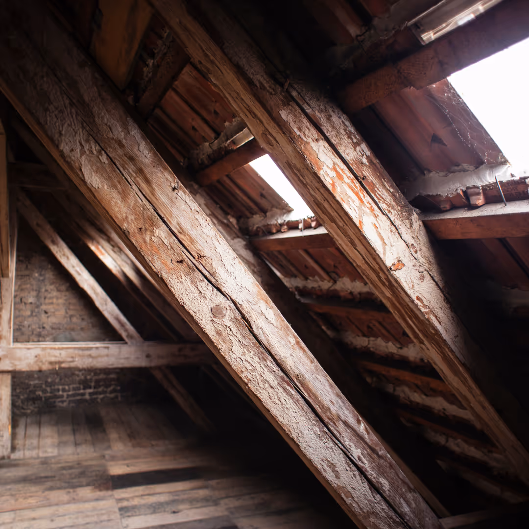 Dachboden mit alten, verwitterten Holzbalken, Holzboden und Dachfenstern, durch die Tageslicht einfällt.