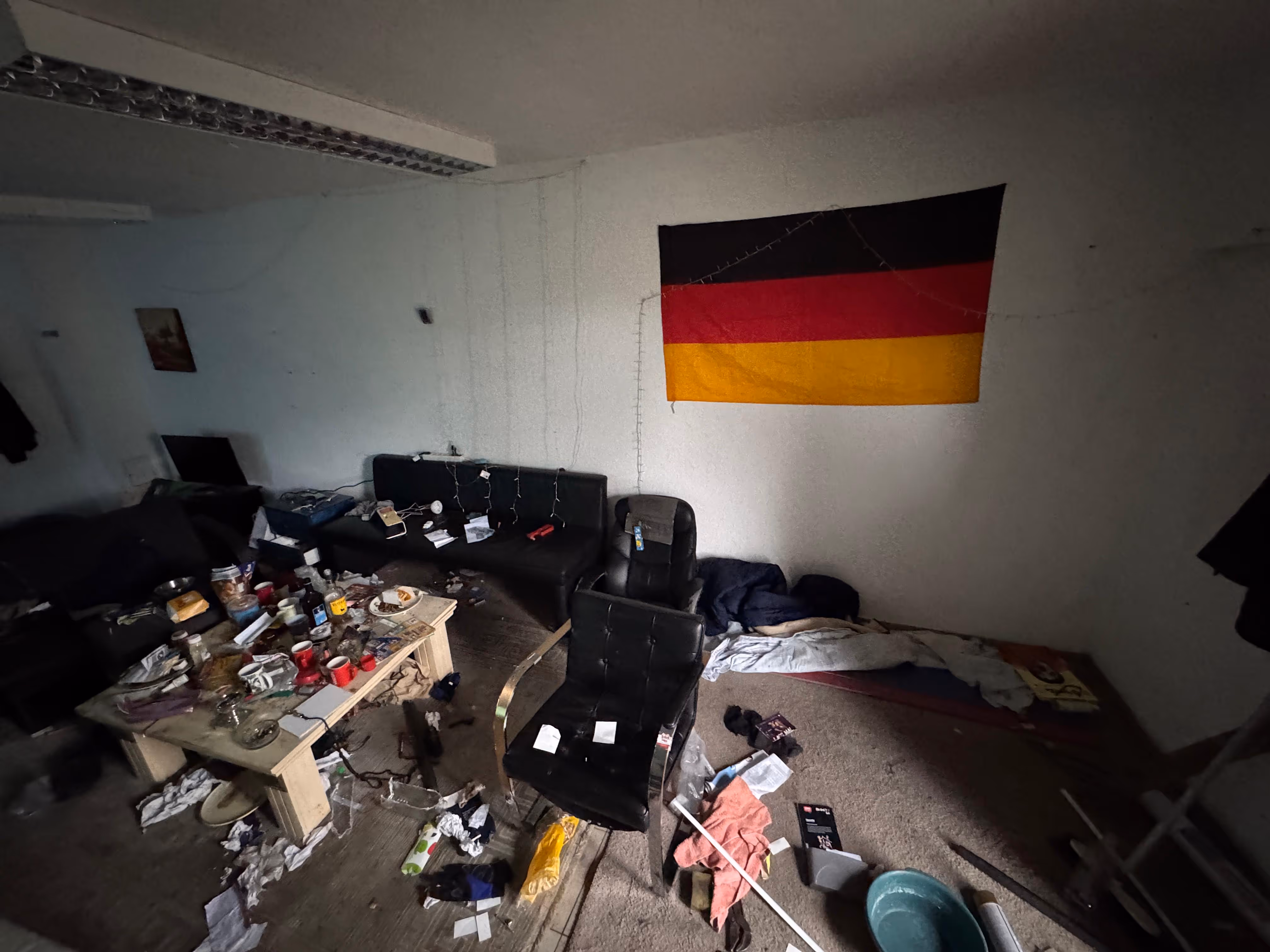 Unordentliches Wohnzimmer mit deutschen Flagge an der Wand, chaotischem Tisch und verstreuten Gegenständen auf dem Boden.