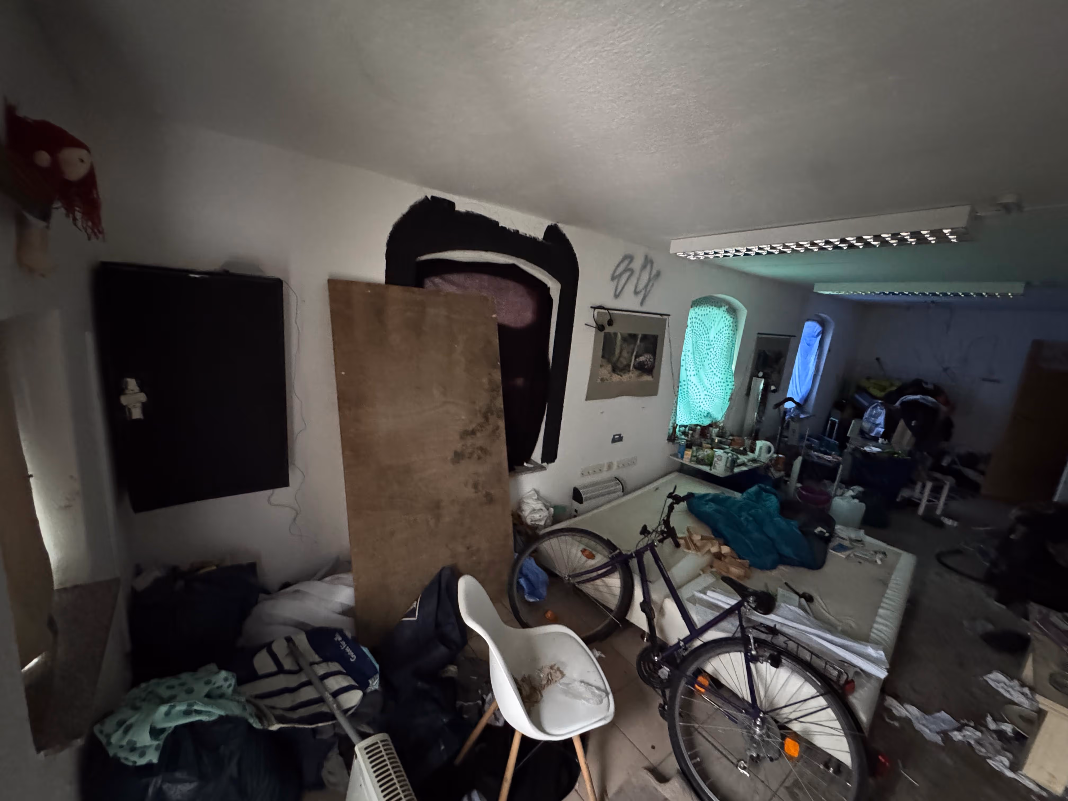 Unordentlicher Raum mit einem Fahrrad, einem weißen Stuhl, verstreuten Gegenständen und einem Bett mit blauer Decke unter Fenstern mit Sichtschutz.
