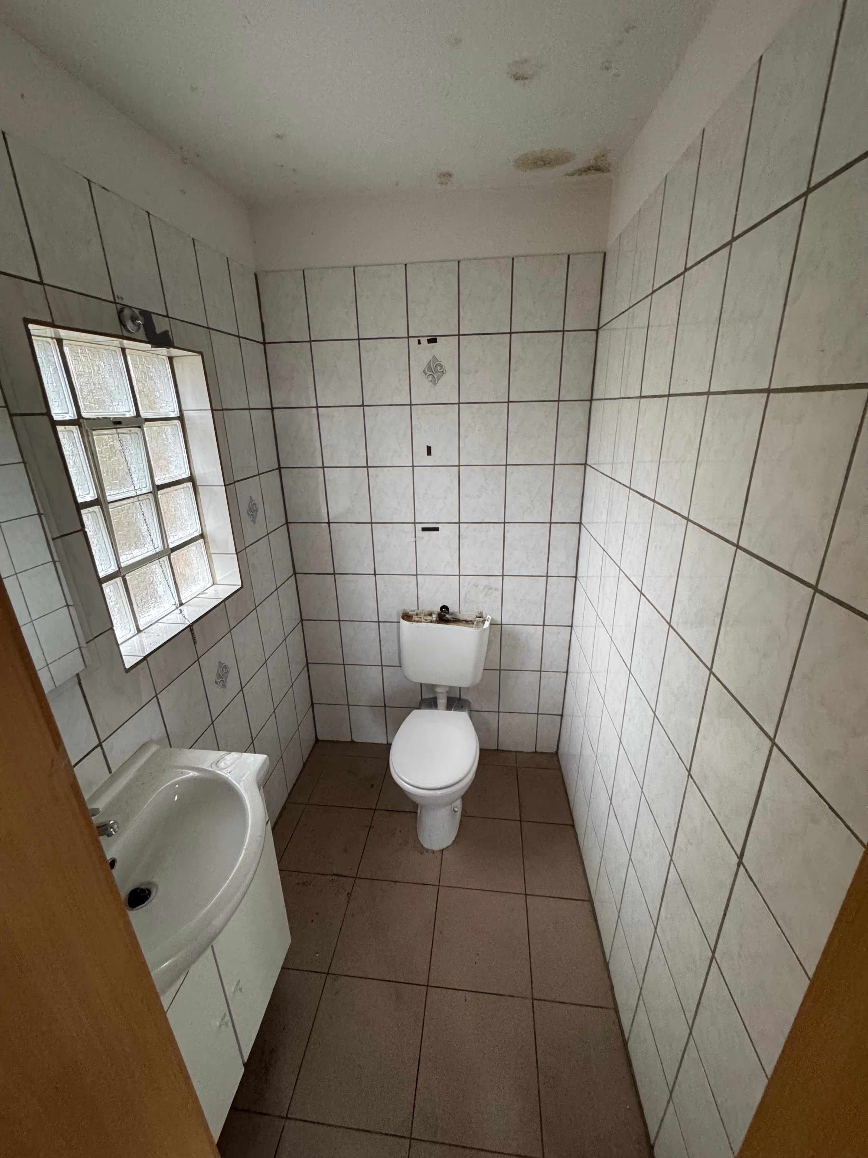 Kleines Badezimmer mit weiß gefliesten Wänden, WC und Waschbecken, Decke mit Feuchtigkeitsschäden.