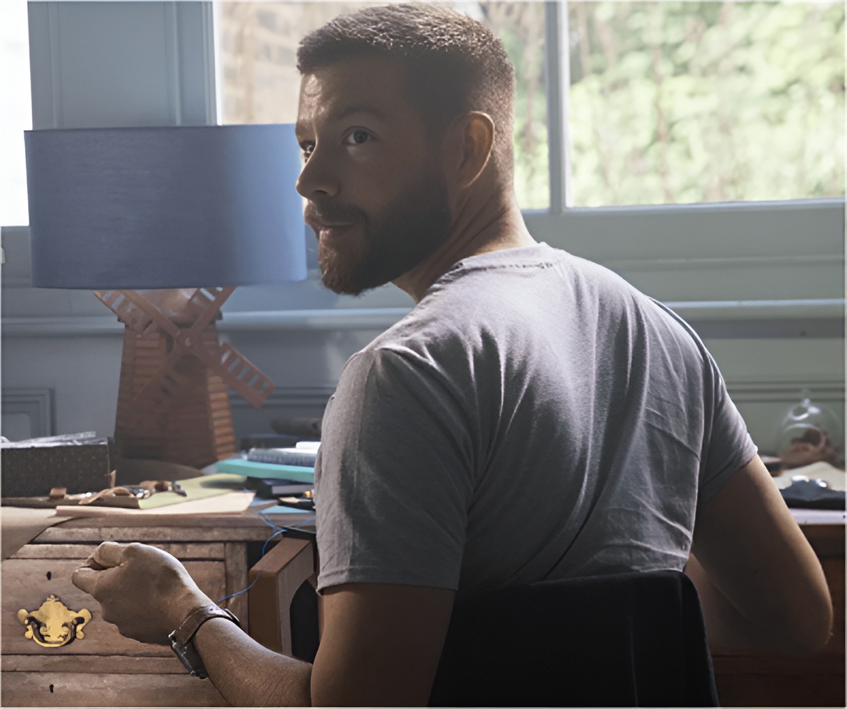 Bearded man in a gray t-shirt sitting at a wooden desk looking back toward the camera.