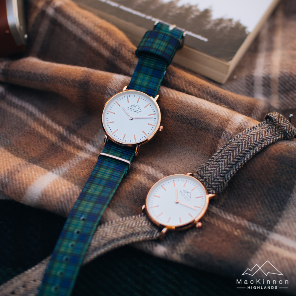 Two elegant wristwatches with white faces and gold rims, one with a blue-green plaid strap and the other with a brown herringbone strap, resting on a warm plaid fabric.