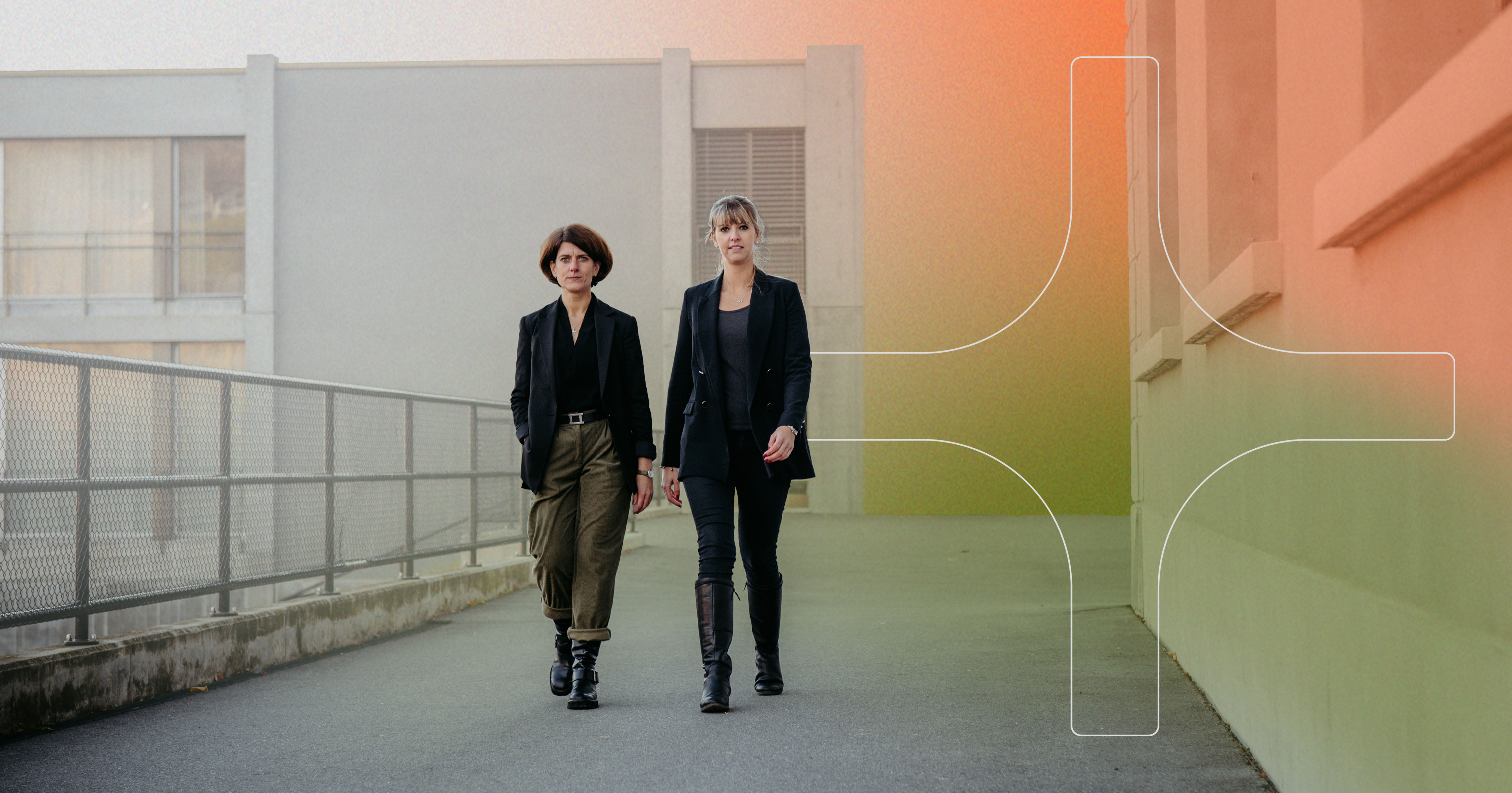 Two women walking confidently on an outdoor path beside a building with a chain-link fence on the left.