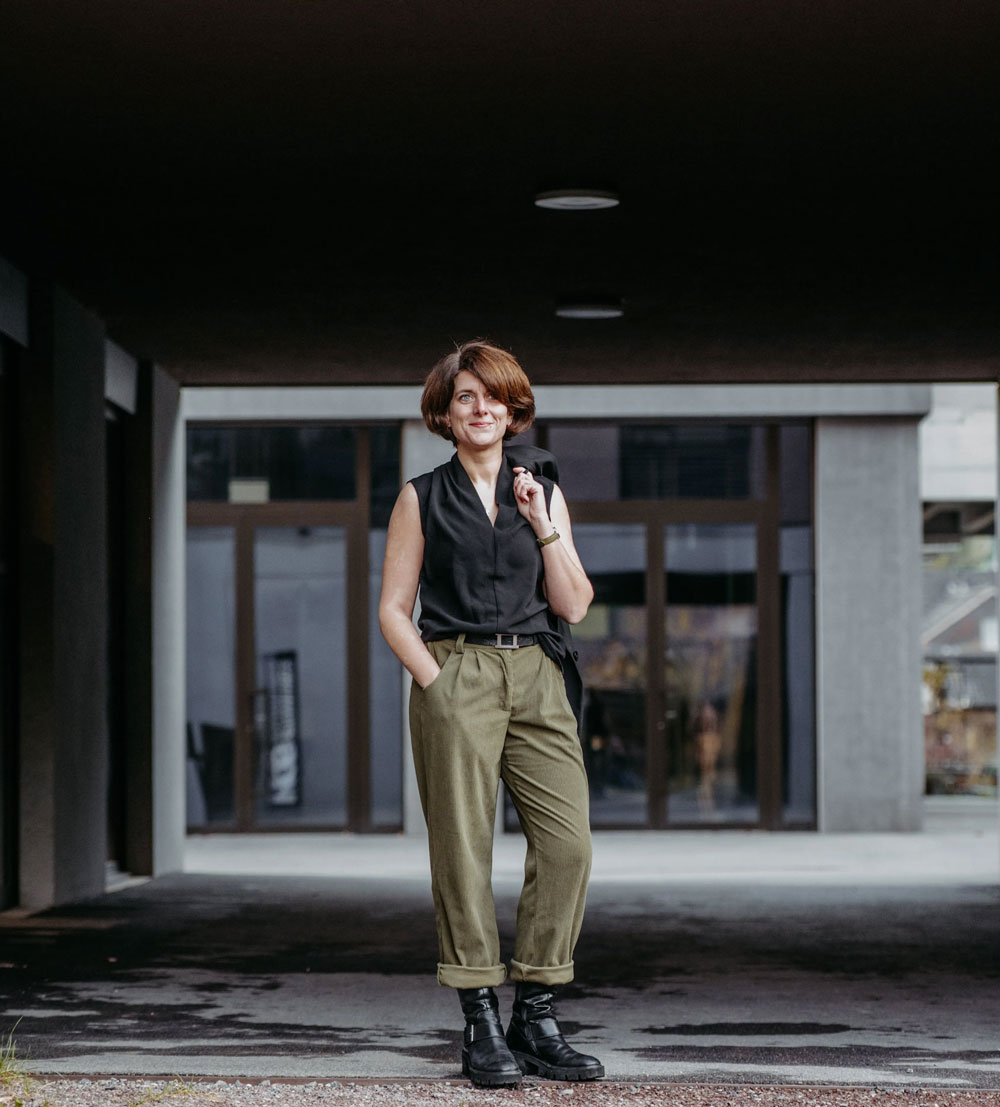 Woman with short brown hair wearing a sleeveless black top, olive green pants, and black boots standing outdoors under a building overhang.