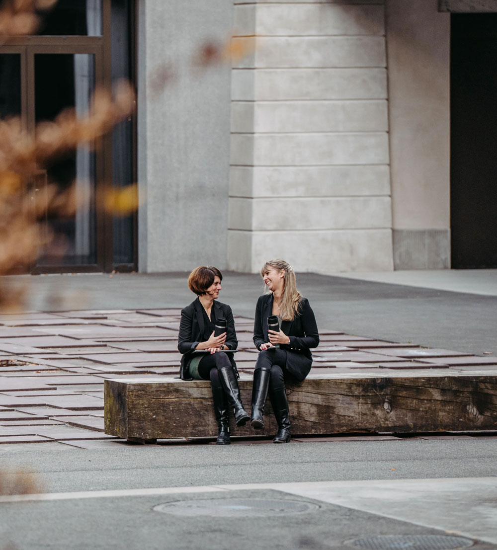 Two women sitting on a large wooden bench outside, each holding a travel coffee mug and smiling at each other.