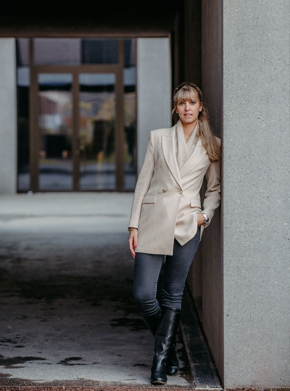 Woman in beige blazer, gray pants, and black boots leaning against a concrete wall near a glass doorway.