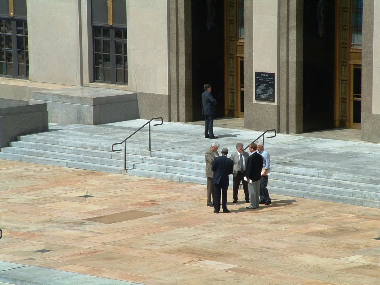 Five men in business attire standing and conversing on a stone plaza in front of a large building entrance with stairs and handrails.