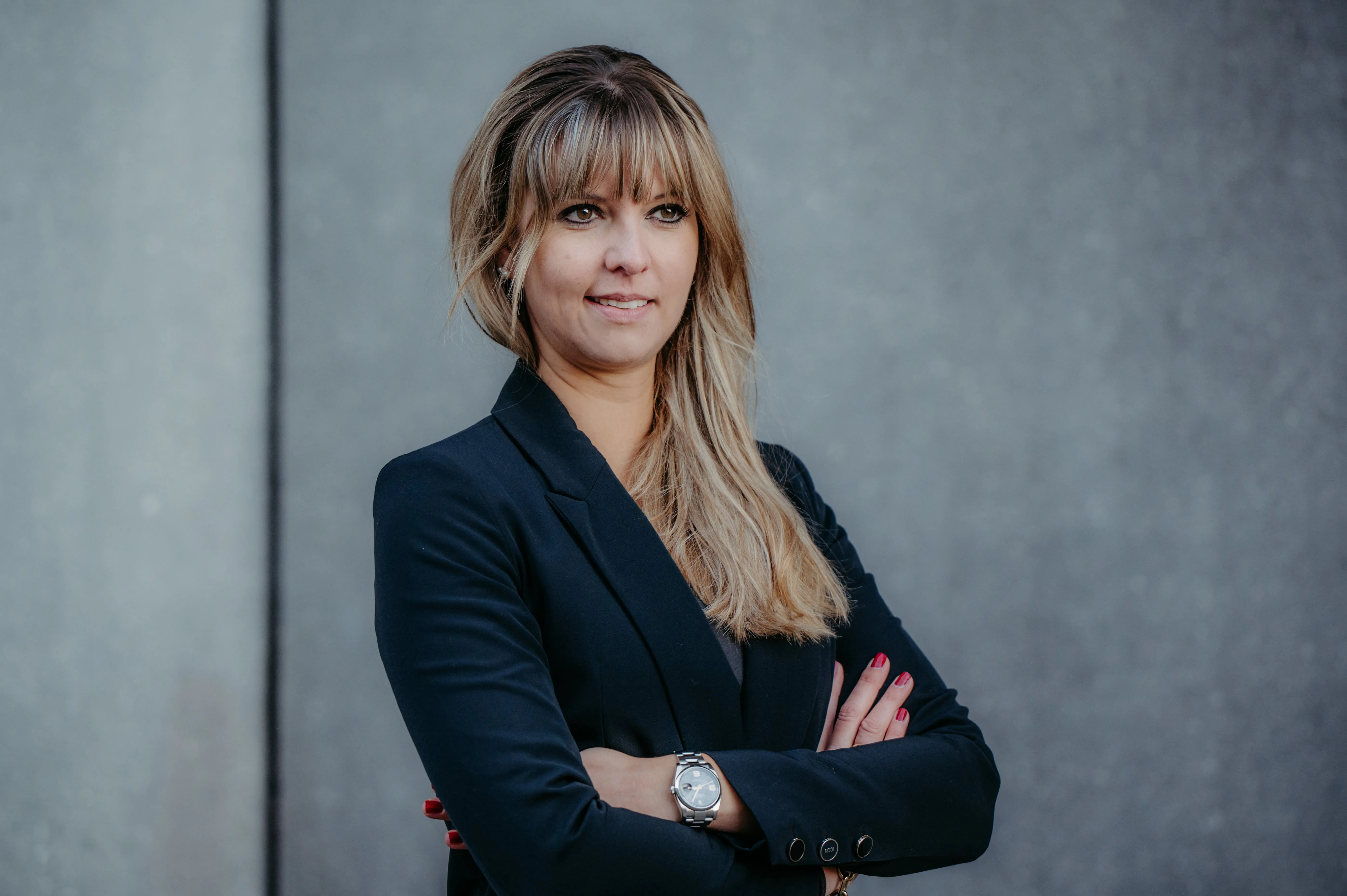 Woman with long blonde hair wearing a black blazer, standing with arms crossed against a gray background.