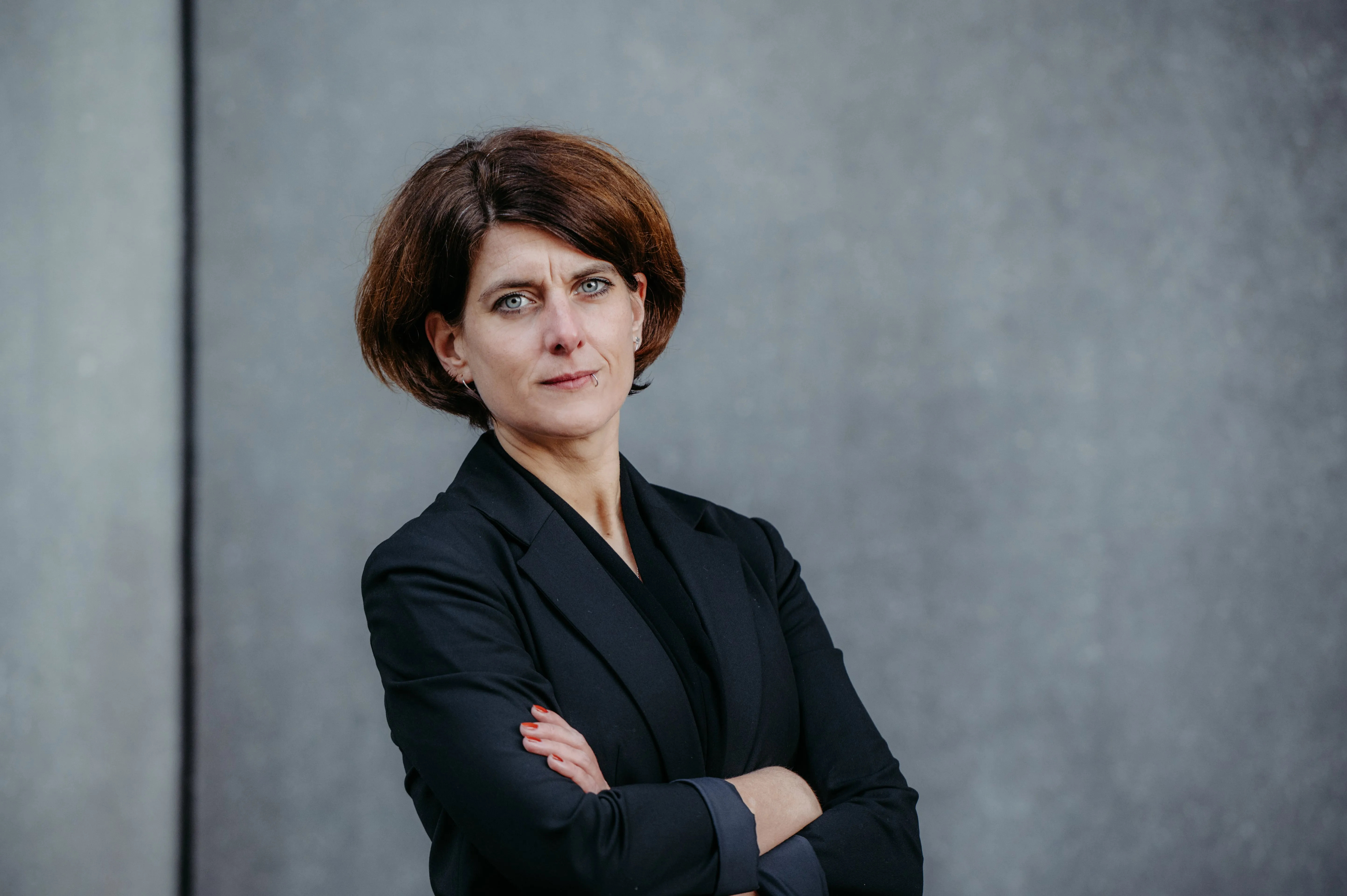 Confident woman with short brown hair wearing a black blazer standing with arms crossed against a gray wall.