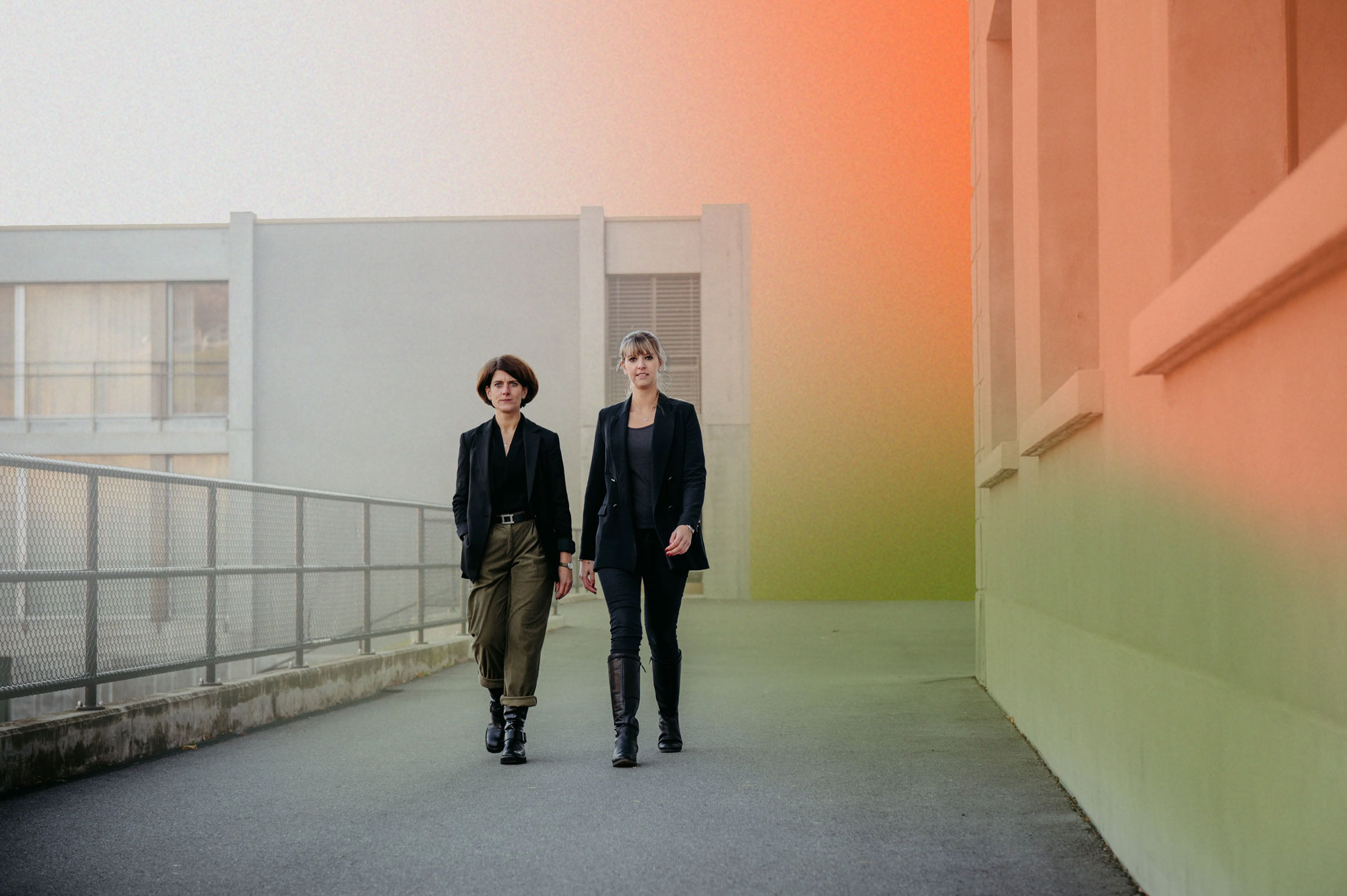 Two women walking side by side on a paved path with buildings and a metal fence in the background.