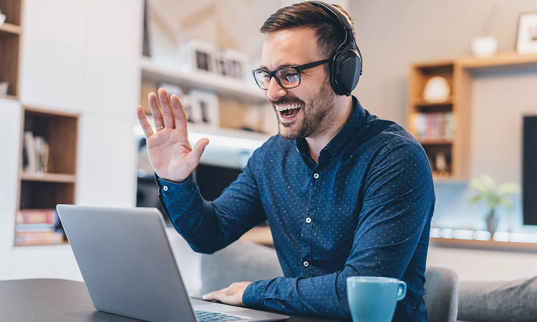 Smiling man wearing glasses and headphones waving during a virtual meeting on his laptop.