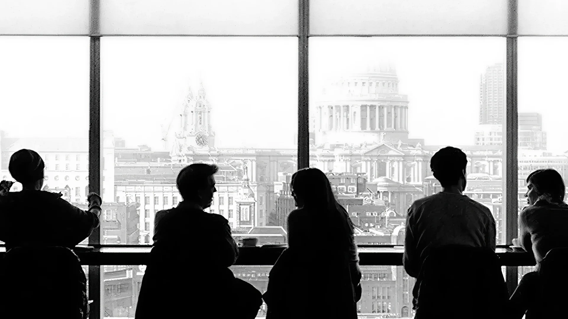 Silhouettes of five people sitting at a counter facing large windows with a cityscape featuring classical architecture in the background.