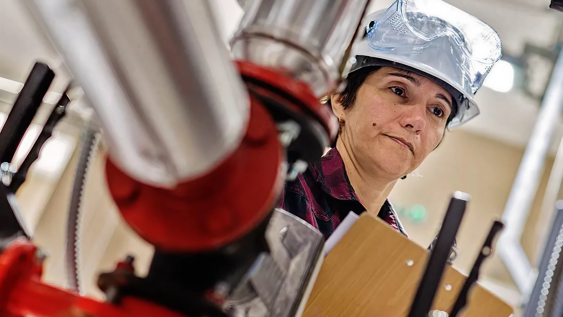 Engineer in protective gear inspecting industrial pipes with a clipboard.