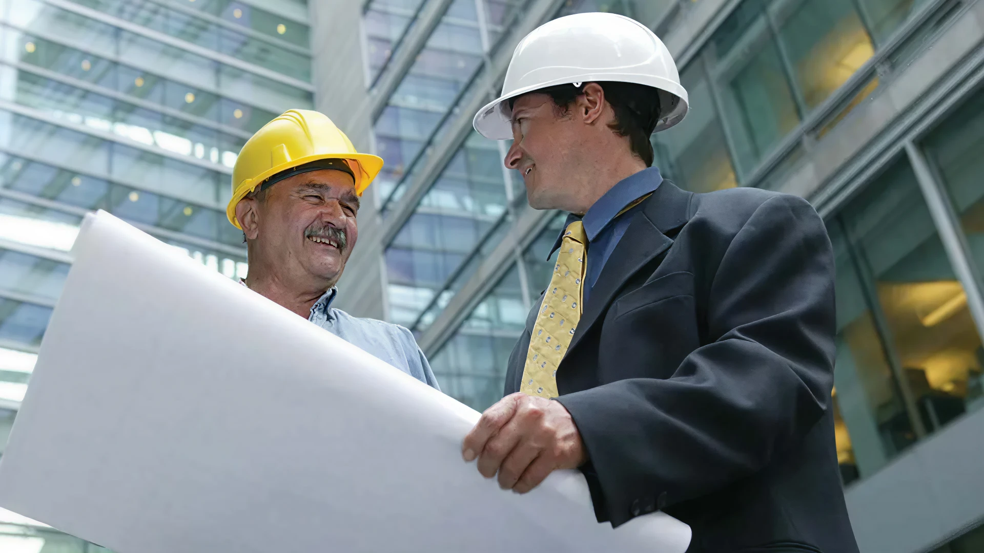 Two workers in hard hats reviewing large building plans outside a modern glass office.