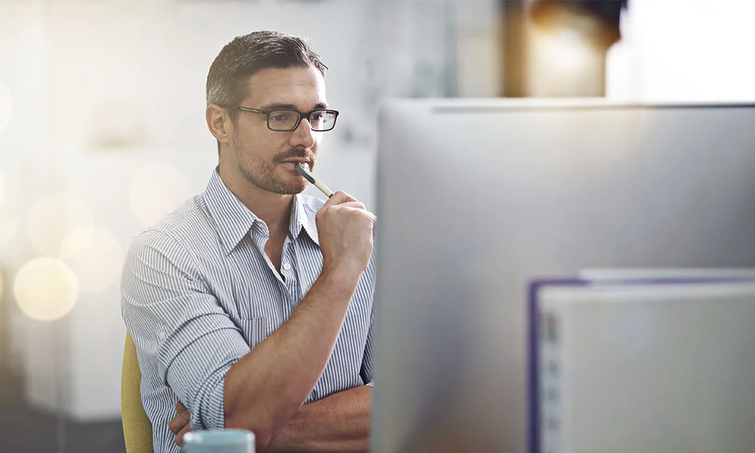 Person sitting at a desk thoughtfully looking at a computer screen while holding a pen.