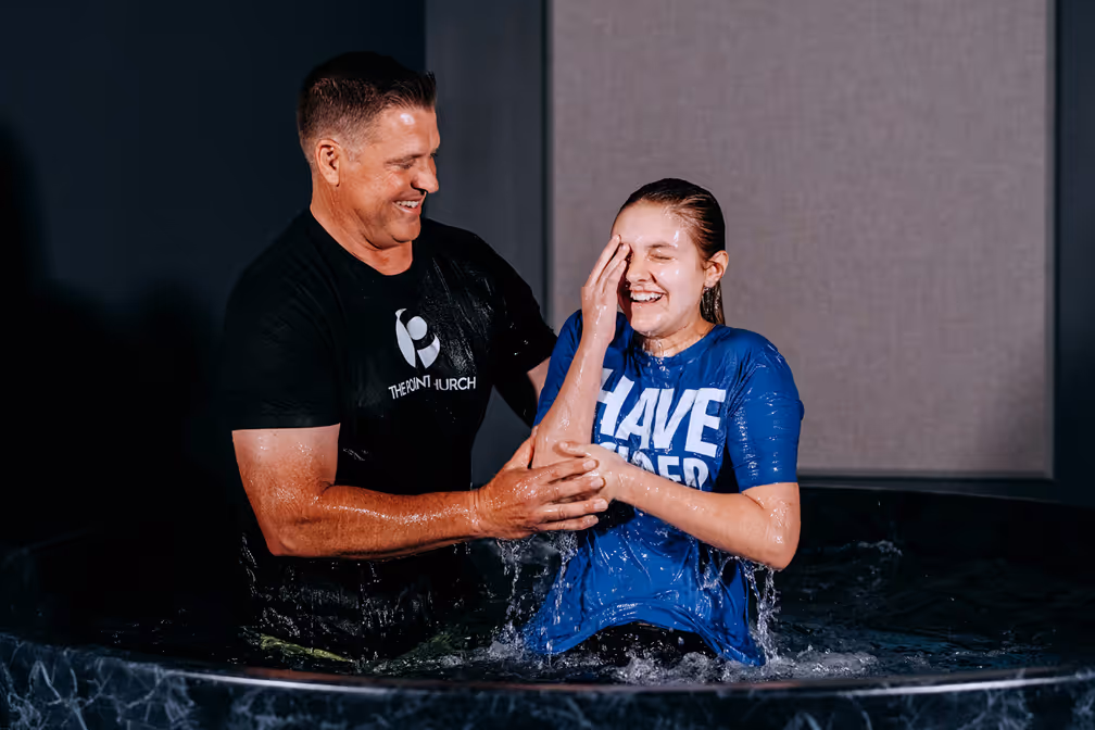 A woman baptizing a young girl in a baptismal pool, with water splashing around them.
