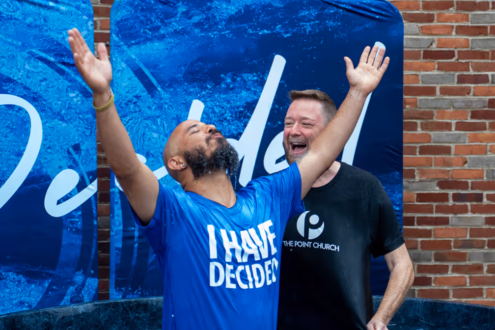 Woman in a blue shirt that says 'I HAVE DECIDED' smiling during a baptism ceremony with two men standing beside her in a church setting.