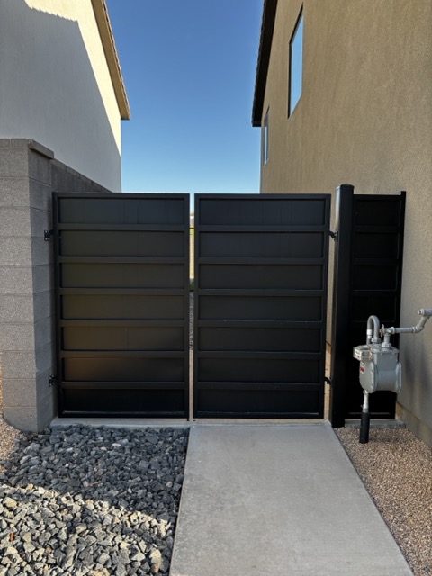 Black metal double gate installed between two buildings with gravel on the left and a gas meter on the right.