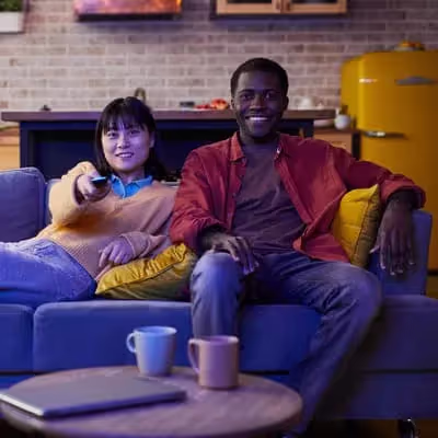 No sofá azul, a mulher deitada e o homem sentado assistem TV, compartilhando risadas e cumplicidade, vivenciando a felicidade em sua sala de estar.