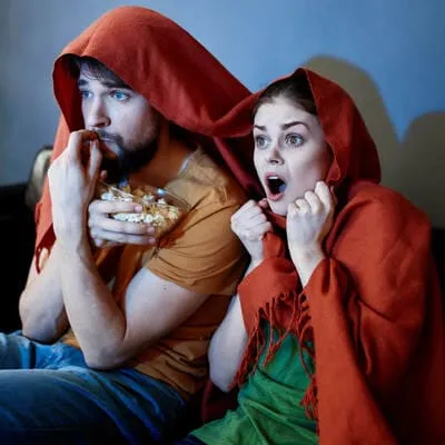 Em meio a um filme de terror, o casal sente o medo tomar conta, com o homem segurando uma tigela de pipoca e a mulher exibindo expressões de surpresa sob uma manta, criando uma atmosfera de tensão e diversão conjunta.