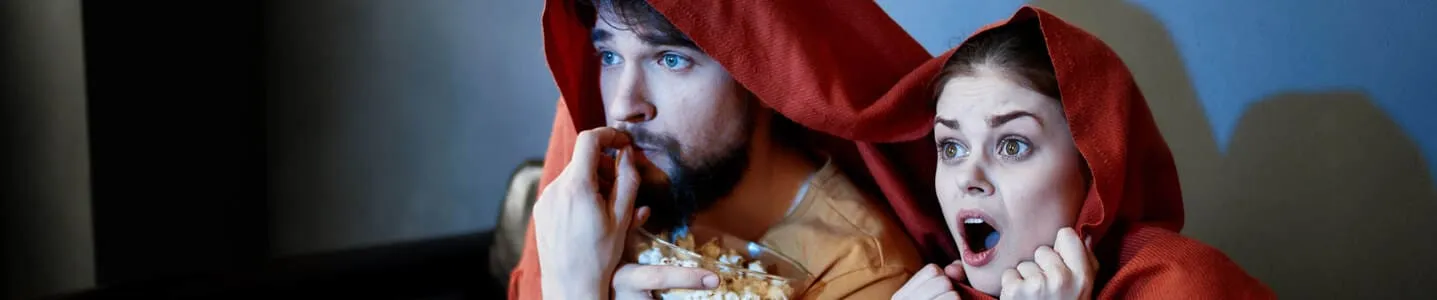 O casal assiste um filme de terror com medo palpável, o homem segura uma tigela de pipoca, enquanto a mulher demonstra surpresa, ambos cobertos por uma manta, compartilhando uma experiência de suspense e entretenimento.