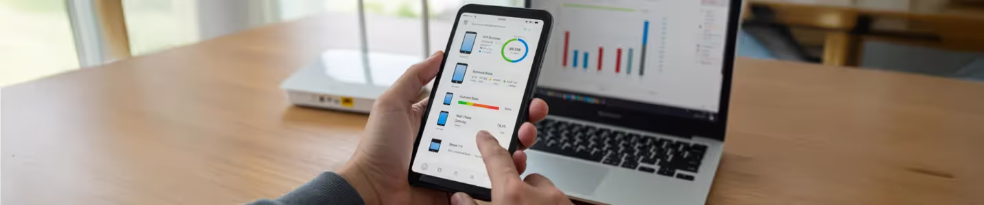 Pessoa usando um smartphone para monitorar o consumo de internet de dispositivos conectados, com um laptop e roteador ao fundo.