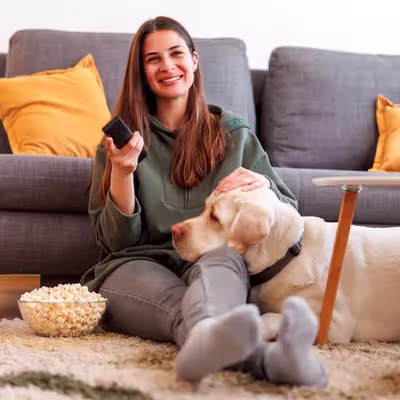 Entre risos, uma jovem mulher se acomoda na sala com pipoca ao lado, controle remoto em mãos, desfrutando de um momento descontraído. Seu companheiro de quatro patas encontra conforto, repousando carinhosamente em seu colo.