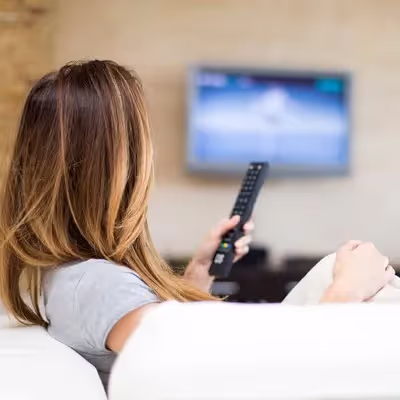 Na sala de estar, a jovem mulher se acomoda no sofá branco, assistindo TV e aproveitando um merecido momento de descontração em sua casa.