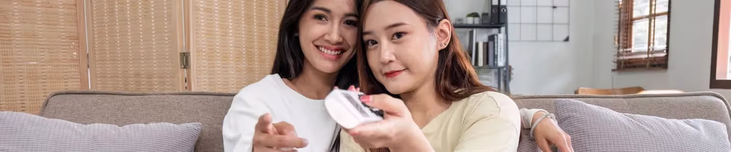 Duas amigas, sintonizadas, apontam o controle remoto para a TV, escolhendo juntas o próximo programa para assistir.