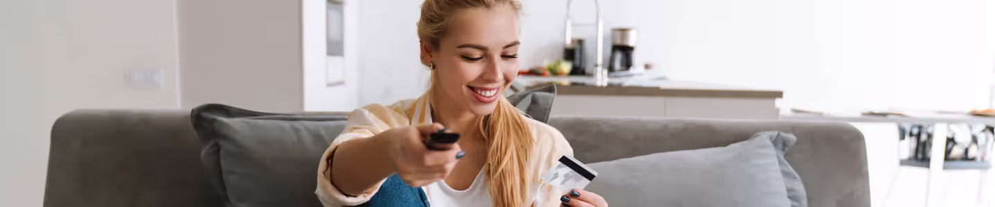 Sentada no sofá, uma jovem mulher segura o controle remoto e o cartão de crédito, feliz por pagar a fatura da Oi TV de forma fácil e conveniente, demonstrando satisfação com a praticidade do processo de pagamento.