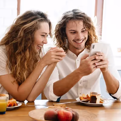 Um casal encantador sorri enquanto utiliza o Oi Fibra em seus smartphones durante o café da manhã, aproveitando a conexão de alta velocidade para iniciar o dia com entusiasmo e praticidade.