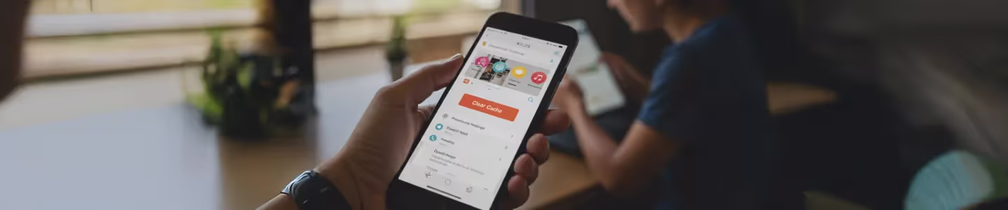 Mão segurando um celular com a opção 'Clear Cache' aberta na tela, enquanto uma pessoa ao fundo usa um tablet.